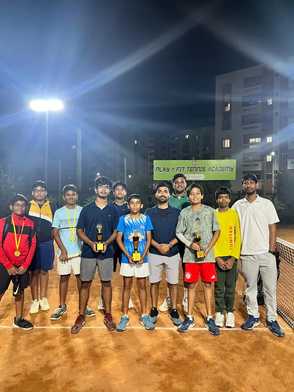 PLAY n FIT juniors and coaches with trophies and medals on court at night under a floodlight, academy banner behind