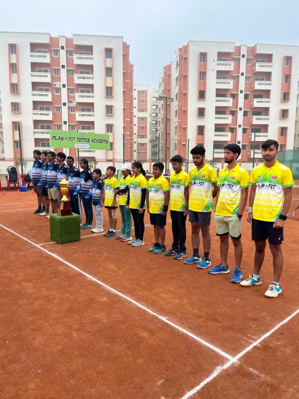 PLAY n FIT players in academy jerseys lined up on the clay court beside a trophy